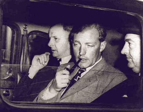 Heath insouciantly smoking his pipe while he is flanked by detectives. Heath was described as the psychopath's psychopath.