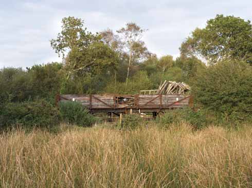 An open wagon with wooden side-boards carrying a cargo of decaying rolling stock frameworks, stands stationary close to Norden station